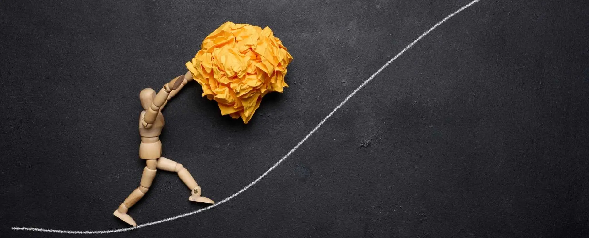 wooden manikin pushing a screwed up ball of paper up a white chalk line hill with a black background